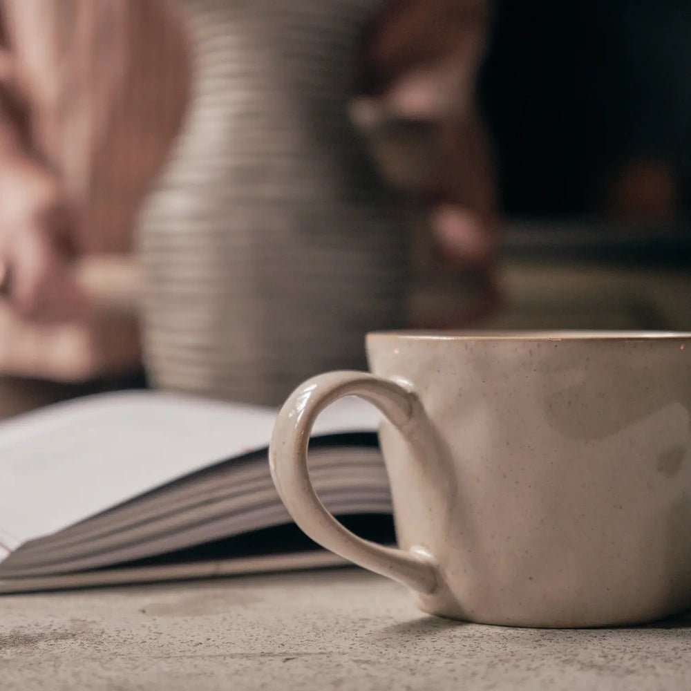 Coleman ceramic mug shown close up with a curved handle, softly uneven cream glaze and gently tapered body, with books and a vase behind.