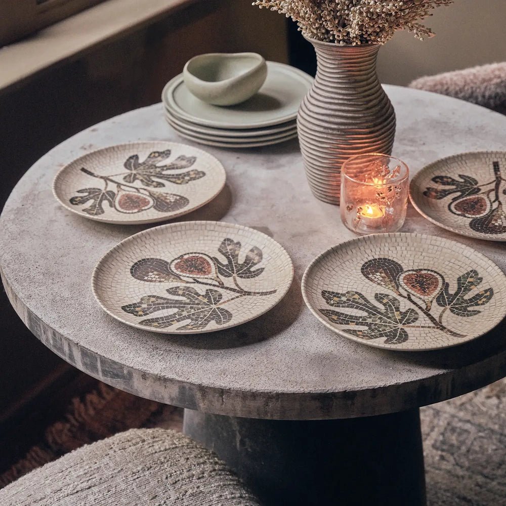 Corbett Mosaic Dinner Plates display fig and leaf designs, arranged around a vase and glowing candle on a round table with stacked bowls.
