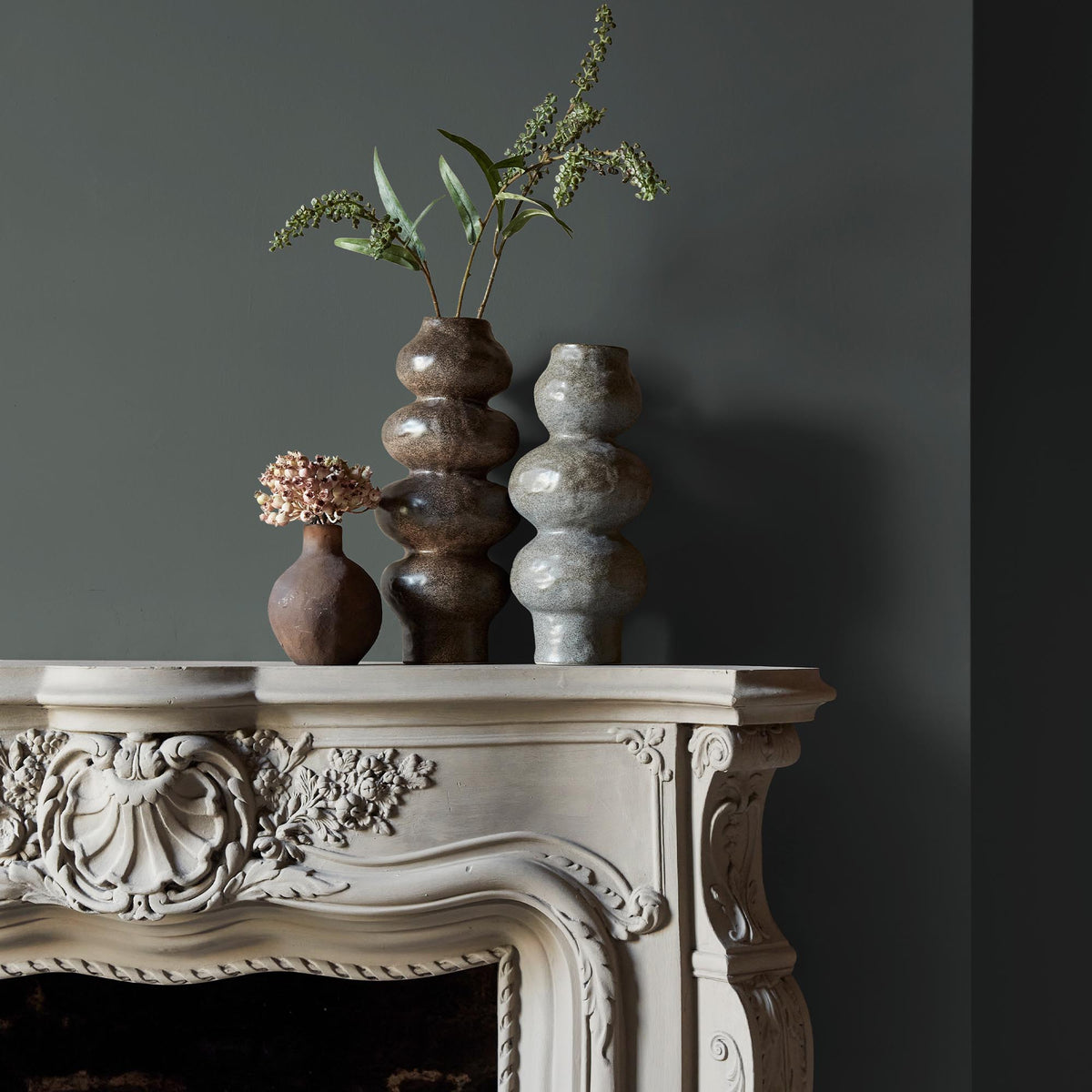 Three ceramic vases sitting on an intricate fireplace mantle in front of a wall in an earthy grey paint colour with green and blue undertones.