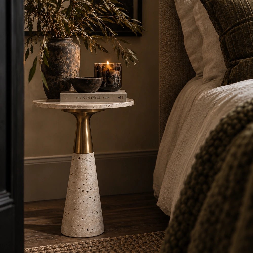 Daytona Marble Side Table with pale stone pedestal and warm metallic top, styled beside a bed with books, candle, bowl and dark decorative vase.