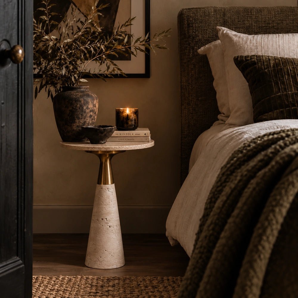 Daytona Marble Side Table with sculptural pale stone base and warm metallic top, styled beside neutral bedding with books, candle, bowl and dark vase.