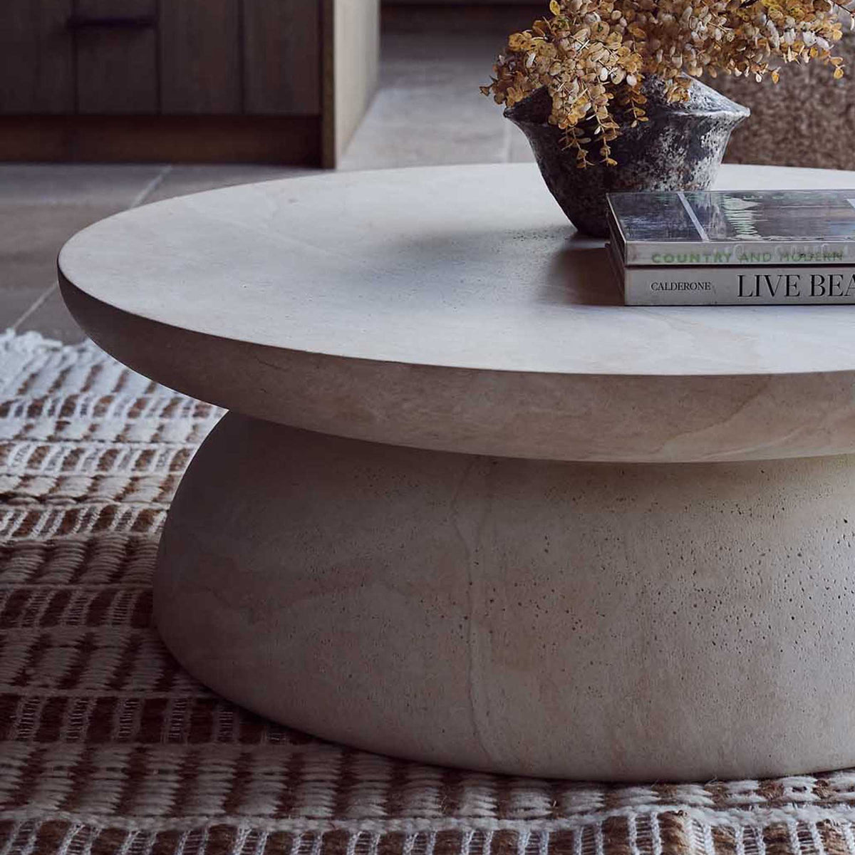 Round travertine effect coffee table with a soft beige finish, subtle cream and grey tones and a sculptural contemporary design. Diablo Round Coffee Table by Abigail Ahern, crafted from glass-reinforced concrete with a textured stone-inspired surface and suitable for both indoor and outdoor spaces. A luxury statement coffee table perfect for modern living rooms and stylish interiors, adding natural texture, timeless elegance and a refined focal point to your home decor.
