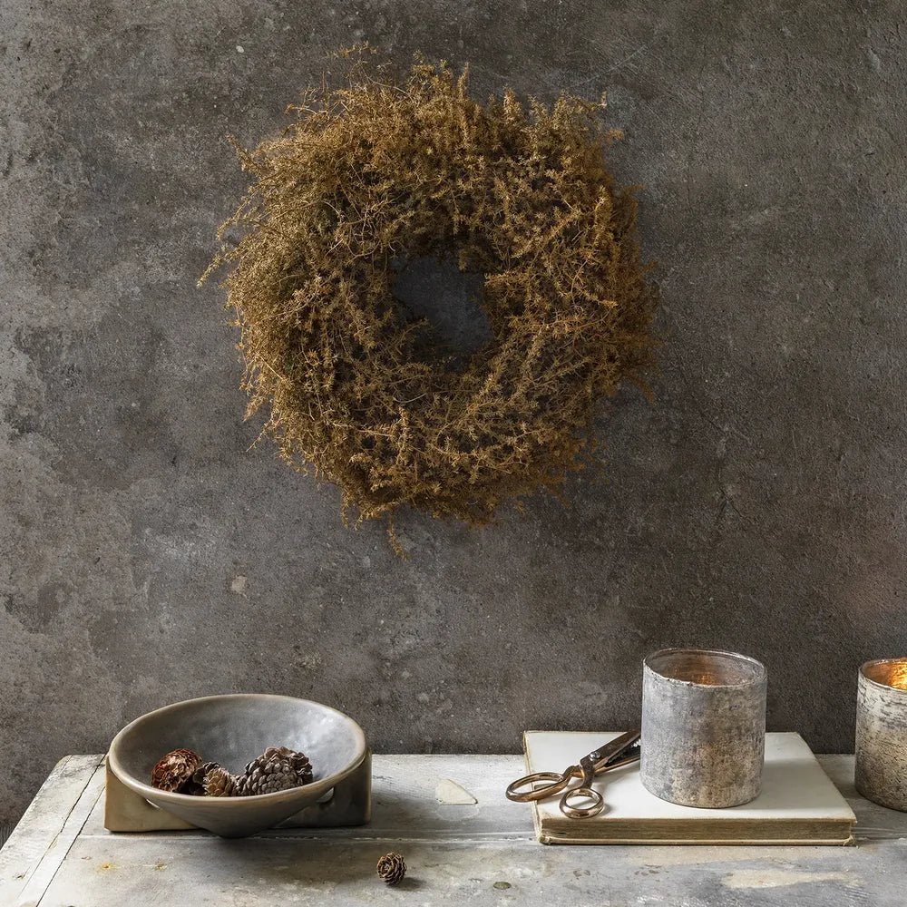 Dried asparagus wreath in russet brown hangs on a textured grey wall above a console table styled with a bowl, scissors and candleholders.