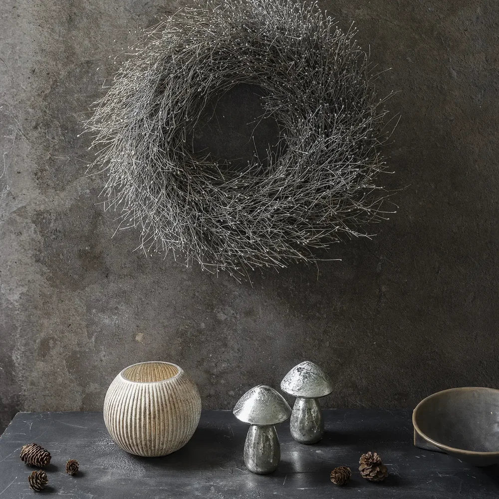 Dried gypsophilia wreath with a round grey twig silhouette hangs on a textured wall above a pale ribbed vase, metallic mushrooms and scattered pine cones.