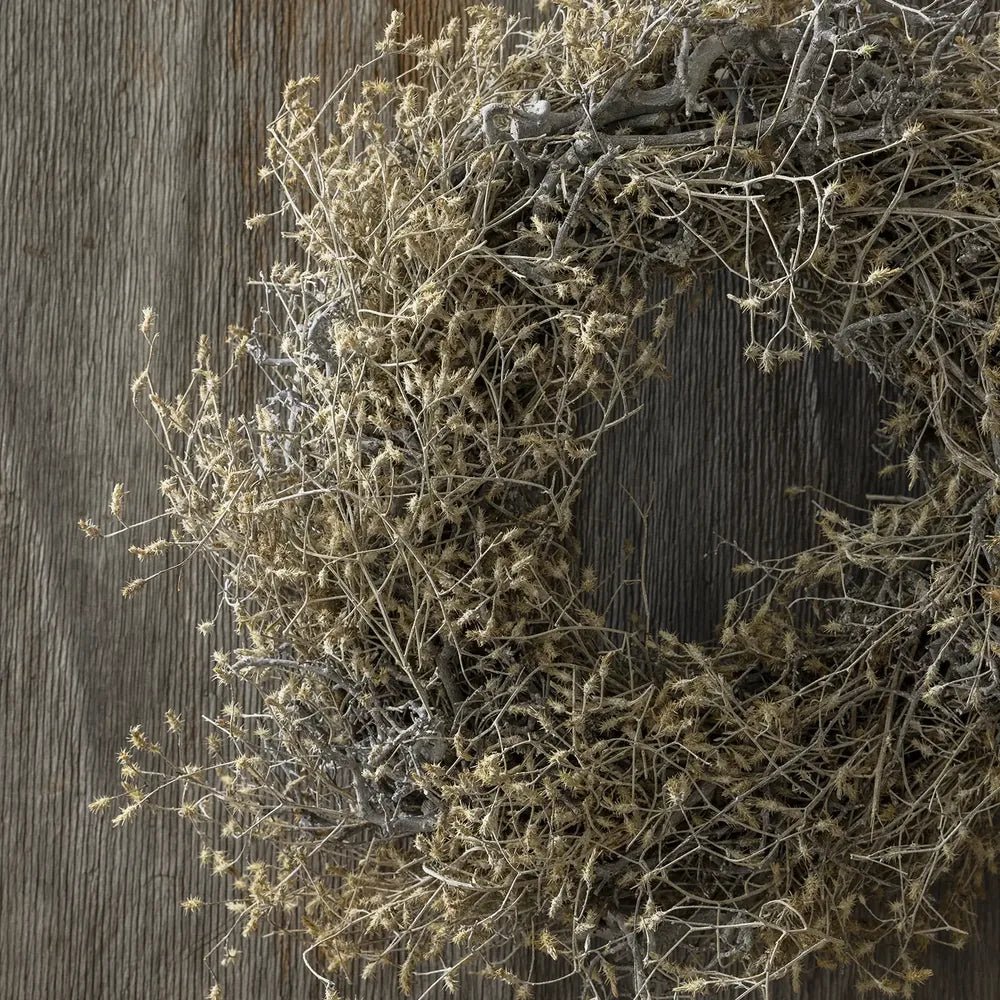 Dried hedgerow wreath displays tangled grey brown stems and scattered dried foliage in close view, with its curved silhouette set against a dark wooden wall.