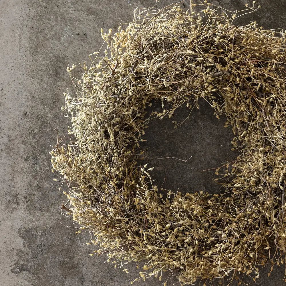 Dried wildwood wreath displays a dense cluster of fine brown branches and dried leaves in close view, highlighting its loosely layered texture against a grey wall.