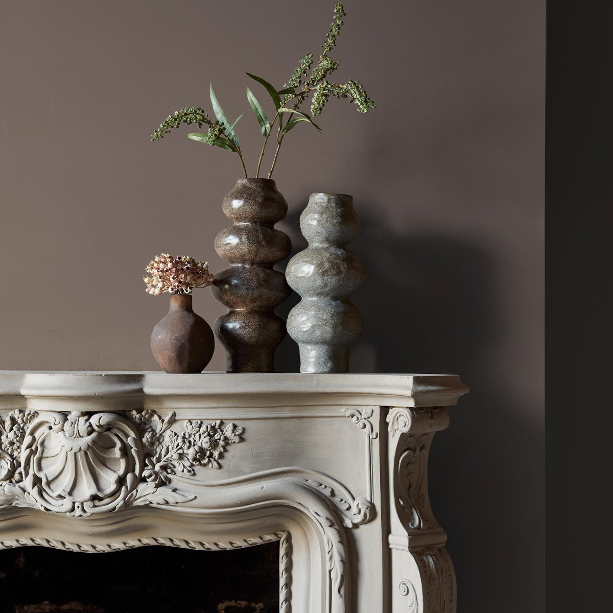 Three ceramic vases sitting on an intricate fireplace mantle in front of a wall in a deep beige paint colour with warm undertones.