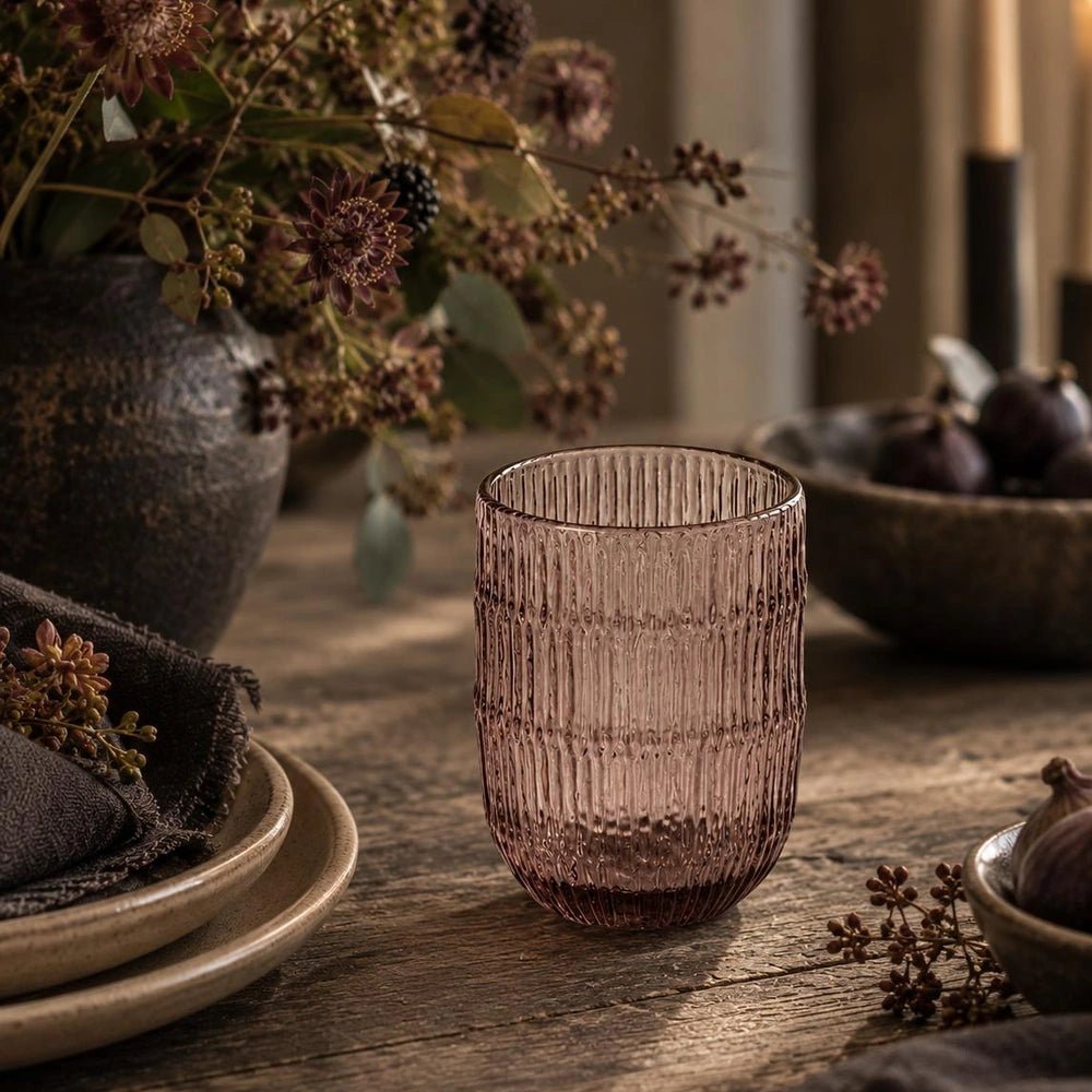 Elden Glass Cup, Blush with ribbed translucent surface, styled on a rustic dining table beside neutral plates, dark florals and softly layered decorative accessories.
