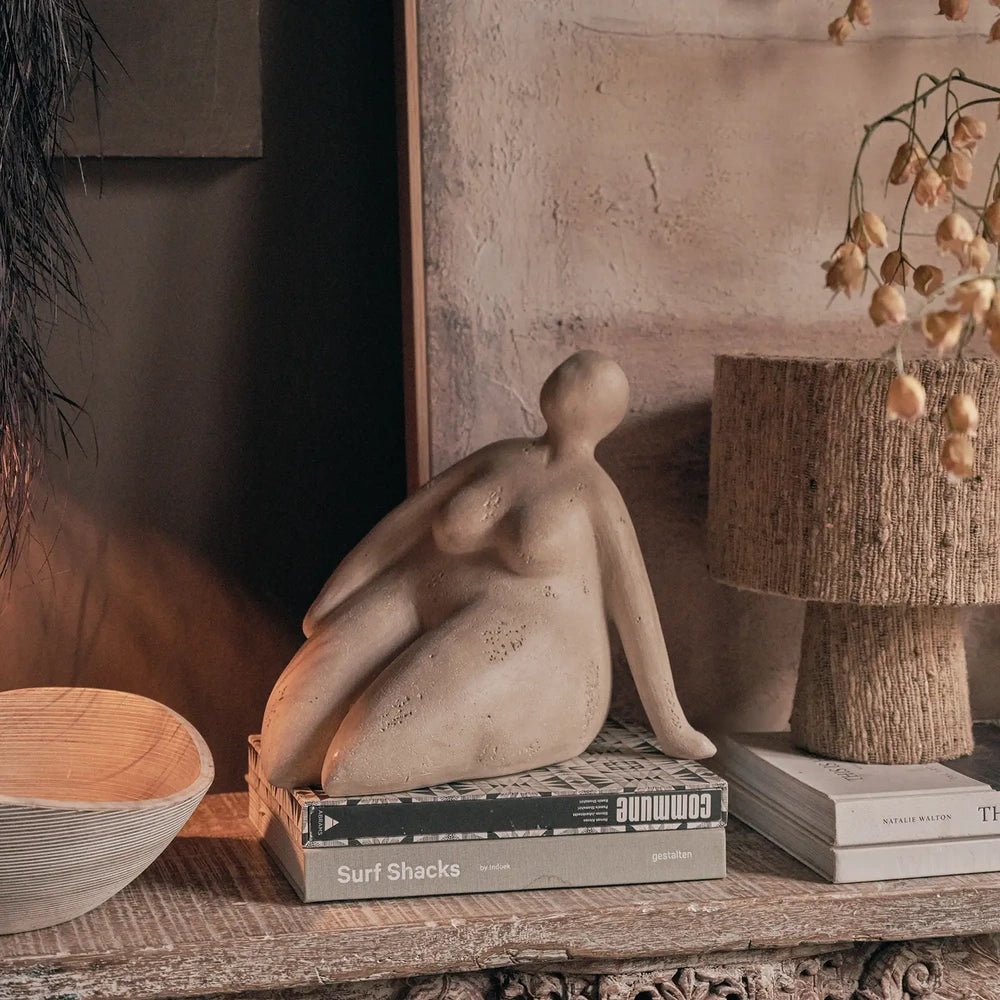 Ellery Figurative Sculpture depicts a seated figure with a smooth neutral finish, displayed on stacked books beside a textured lamp and decorative bowl.