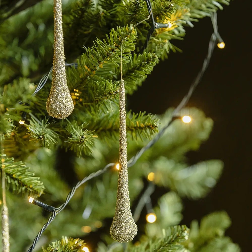 Enzo glitter droplet baubles hang at different heights among green tree branches, with textured pale gold surfaces catching light from a string of warm bulbs.