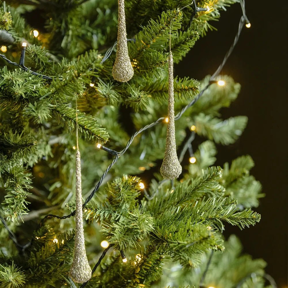 Enzo glitter droplet baubles with elongated tapered shapes and pale gold textured surfaces hang among green Christmas tree branches threaded with warm white lights.