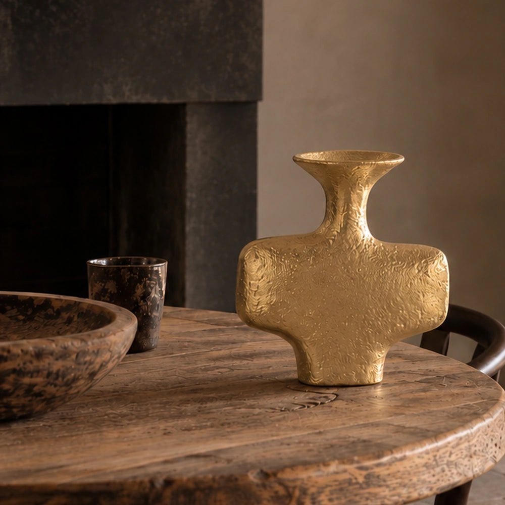 Everley Metal Vase in textured gold finish displayed empty on a rustic wooden dining table beside a dark candle holder and decorative bowl.