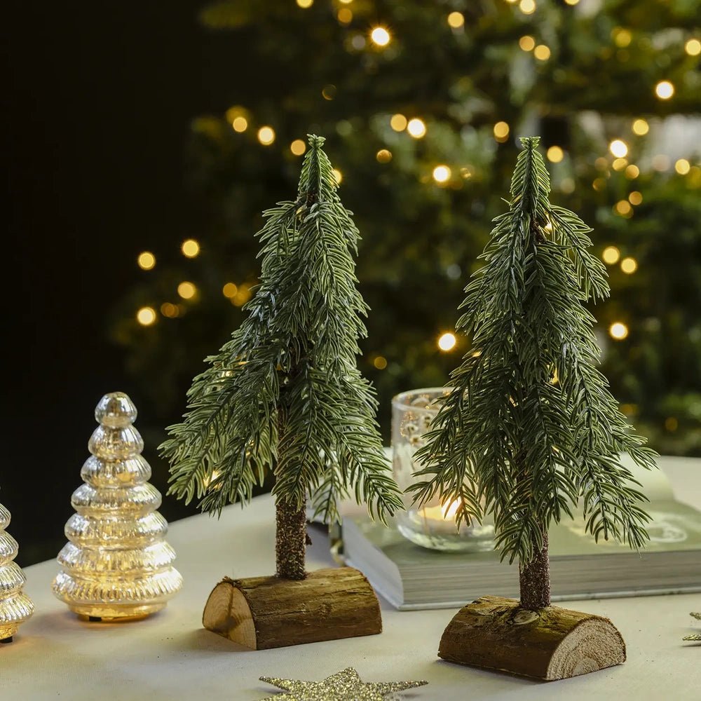 Festive tree decorations form a pair of small green trees on curved wooden bases, displayed beside glowing tree lamps and candleholders against Christmas lights.