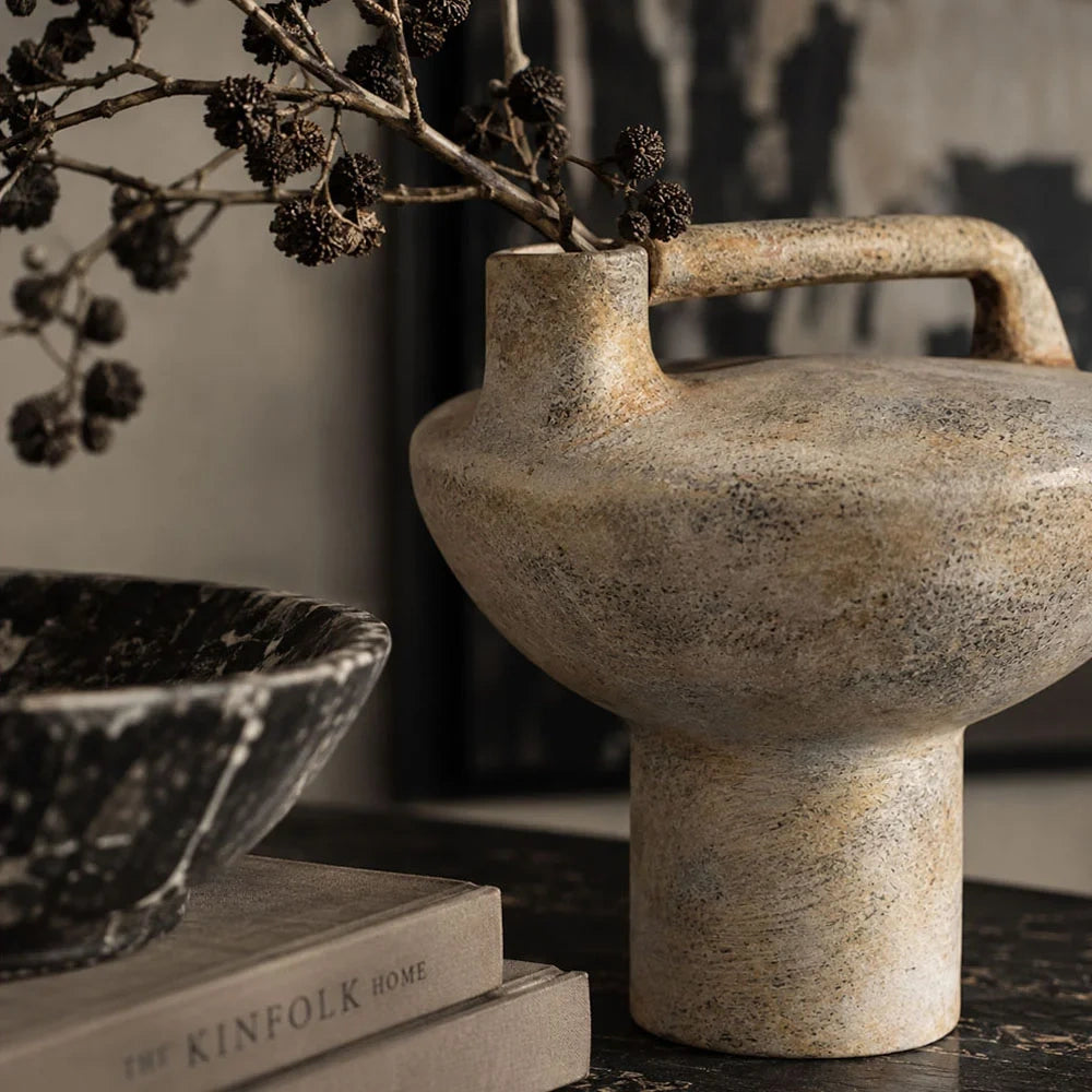 Forbes sand ceramic vase with a rounded bowl shaped body, cylindrical pedestal and raised rectangular handle holds dark branches beside books on a console.