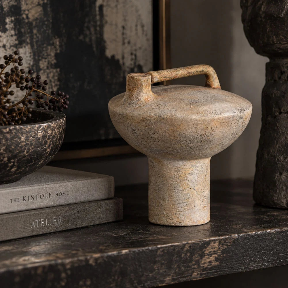 Forbes sand ceramic vase with a textured beige surface, rounded body and rectangular top handle stands on a dark console beside books and a bowl.
