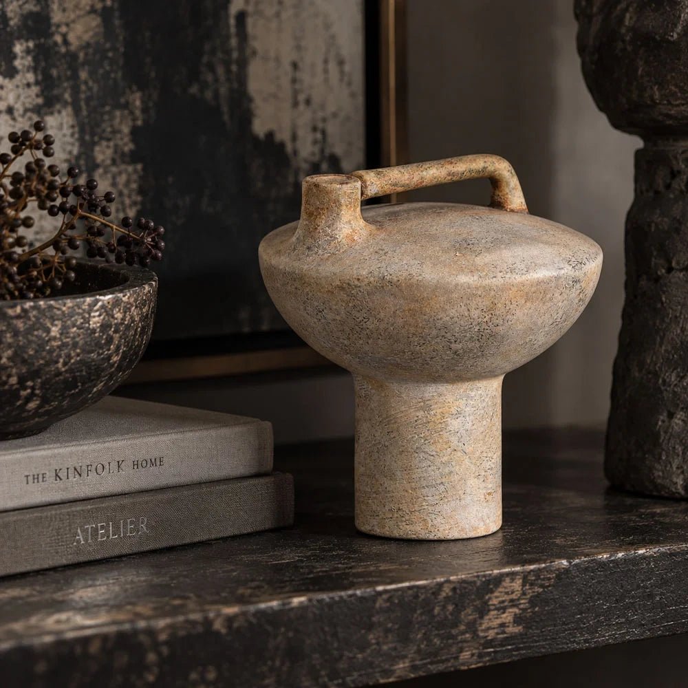 Forbes sand ceramic vase with a textured beige surface, rounded body and rectangular top handle stands on a dark console beside books and a bowl.