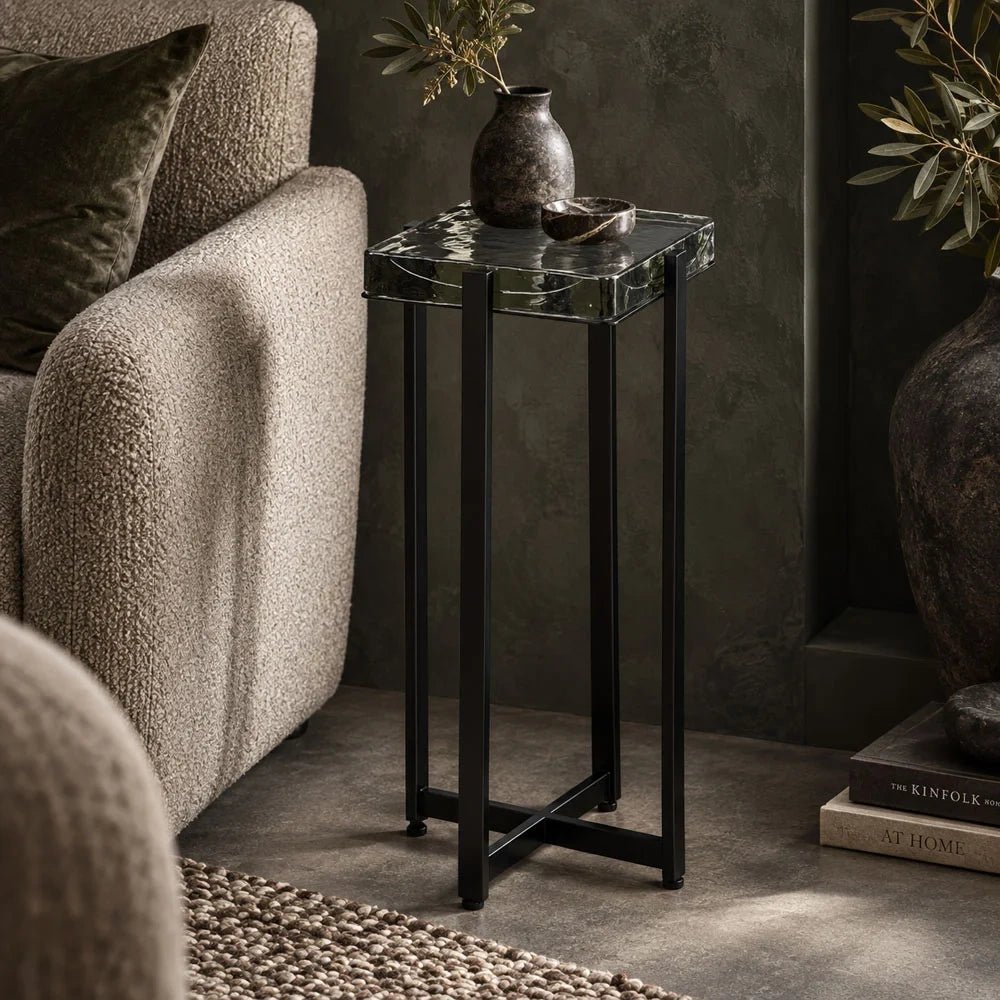 Fordham Glass Side Table features a dark slender frame and square glass top, styled with a vase beside a pale upholstered lounge chair.