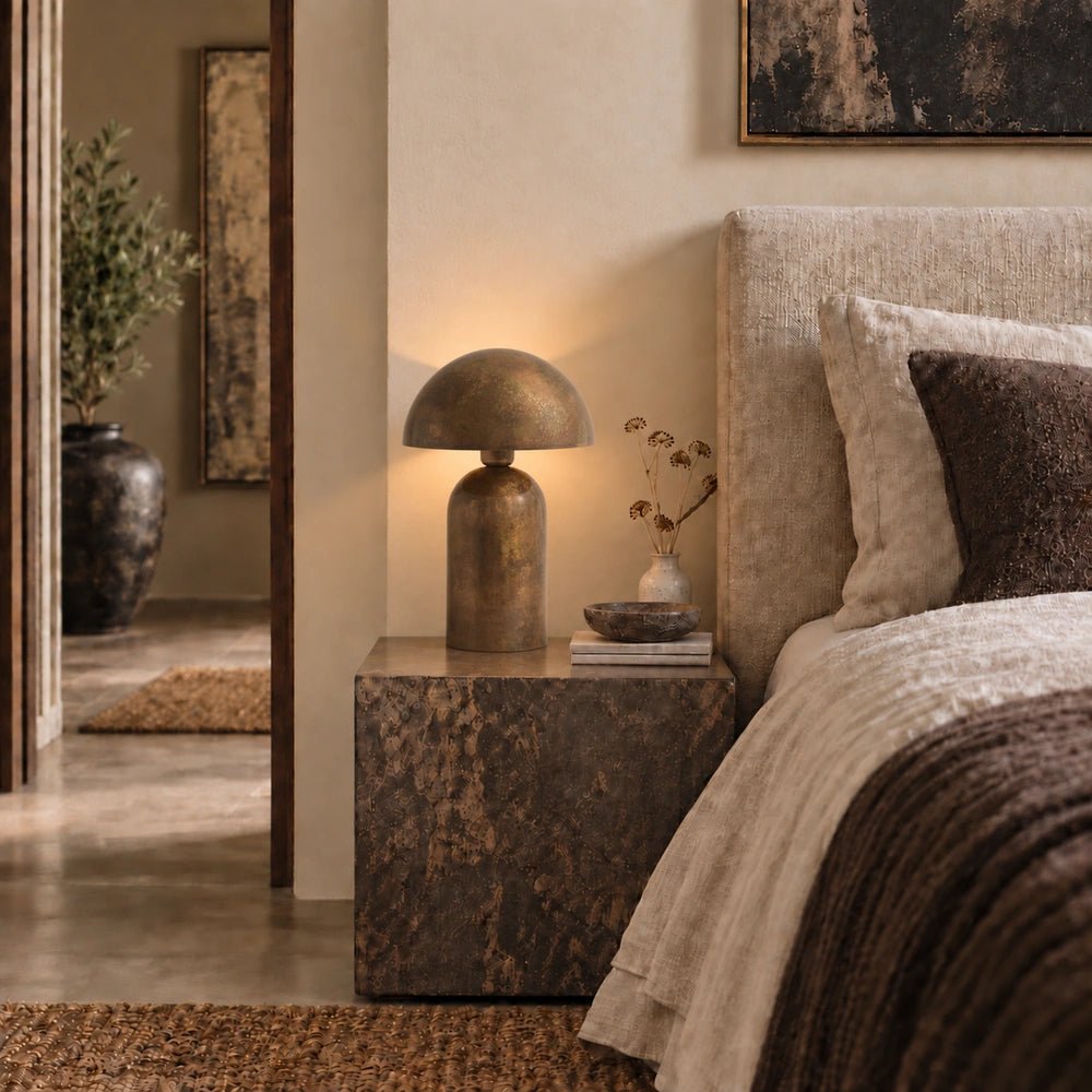 Foxley Metal Table Lamp with dark sculptural metal base and rounded shade, glowing on a bedside table beside layered neutral bedding, artwork and decorative objects.