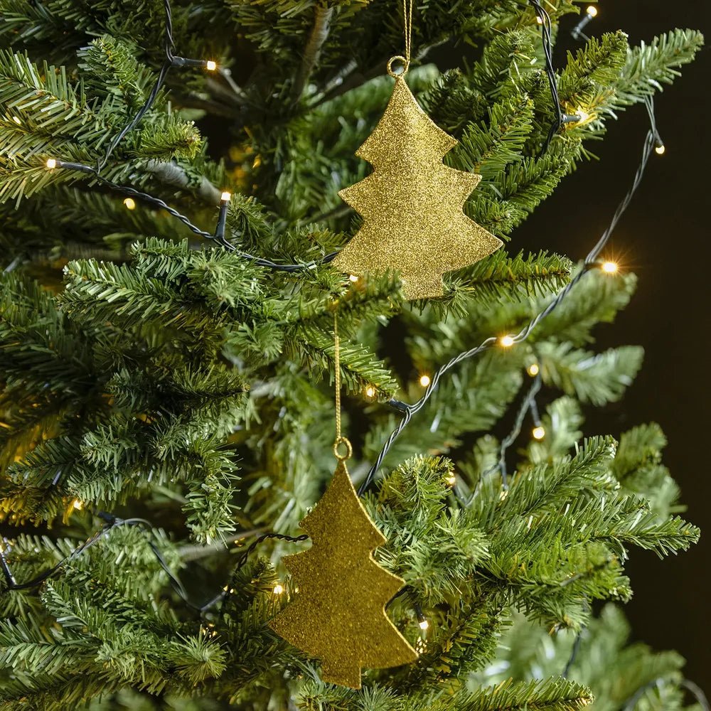 Glittering tree baubles with flat gold silhouettes and sparkling textured surfaces hang among green Christmas tree branches decorated with a string of warm lights.