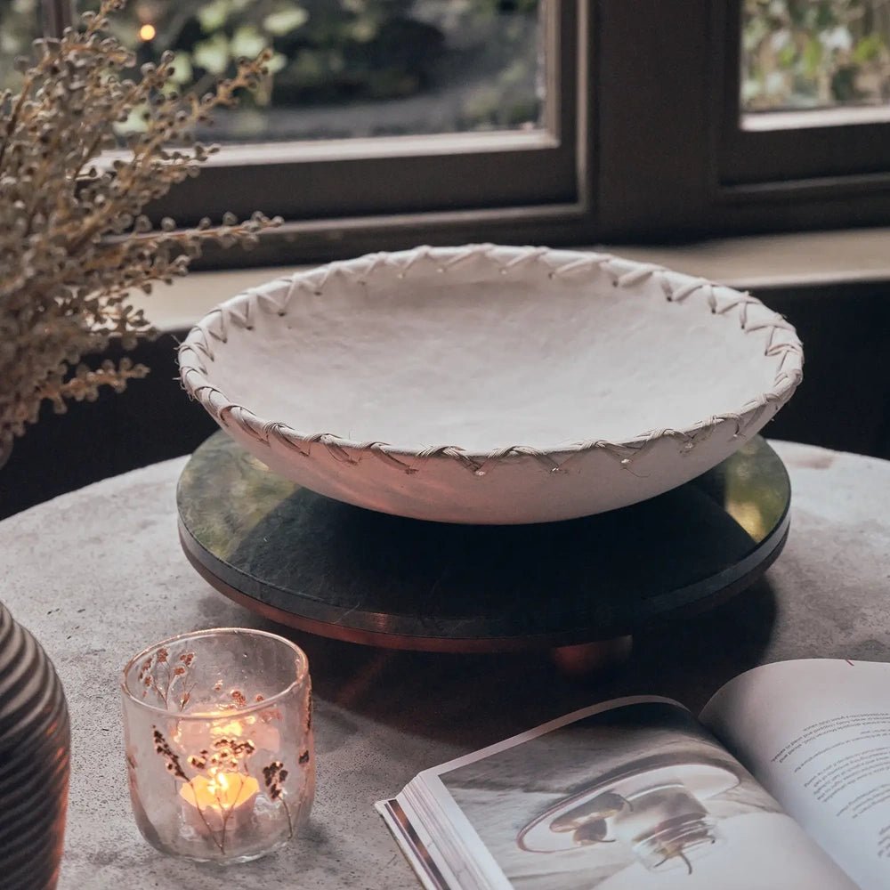Halstead paper mache decorative bowl rests on a dark raised stand beside a window, with its pale shallow interior and laced rim near books and candlelight.