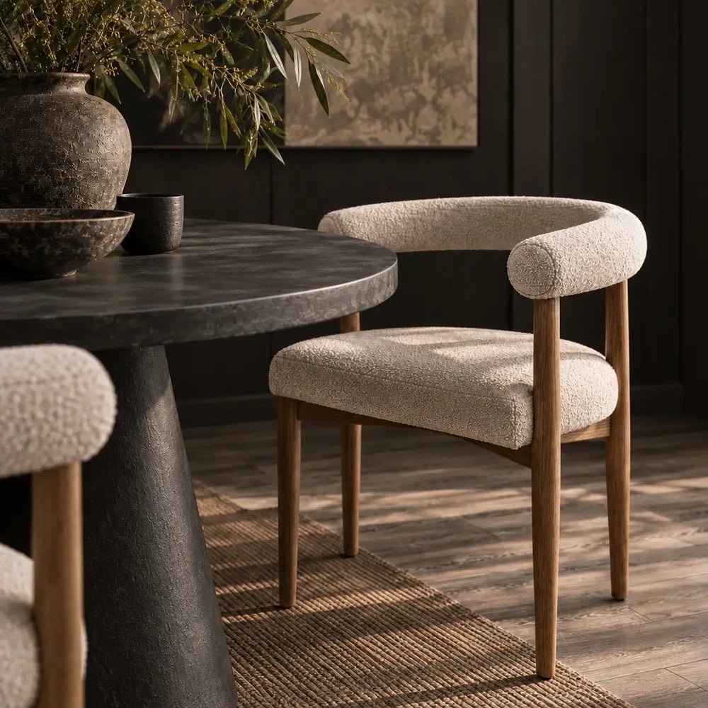 Harrington Boucle Dining Chair features cream textured upholstery, a curved open back and wooden legs, styled beside a round dark dining table.