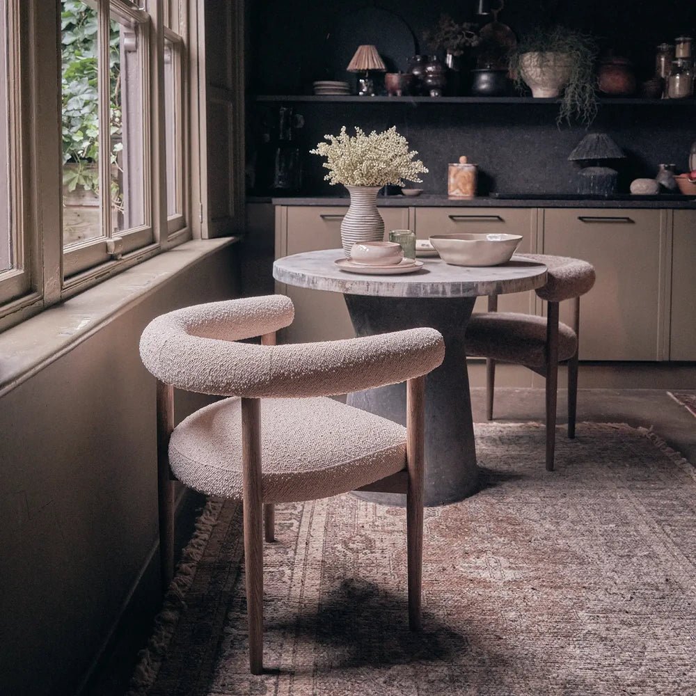 Harrington Boucle Dining Chair features pale textured upholstery and wooden legs, arranged at a round dining table in a kitchen with dark cabinetry.