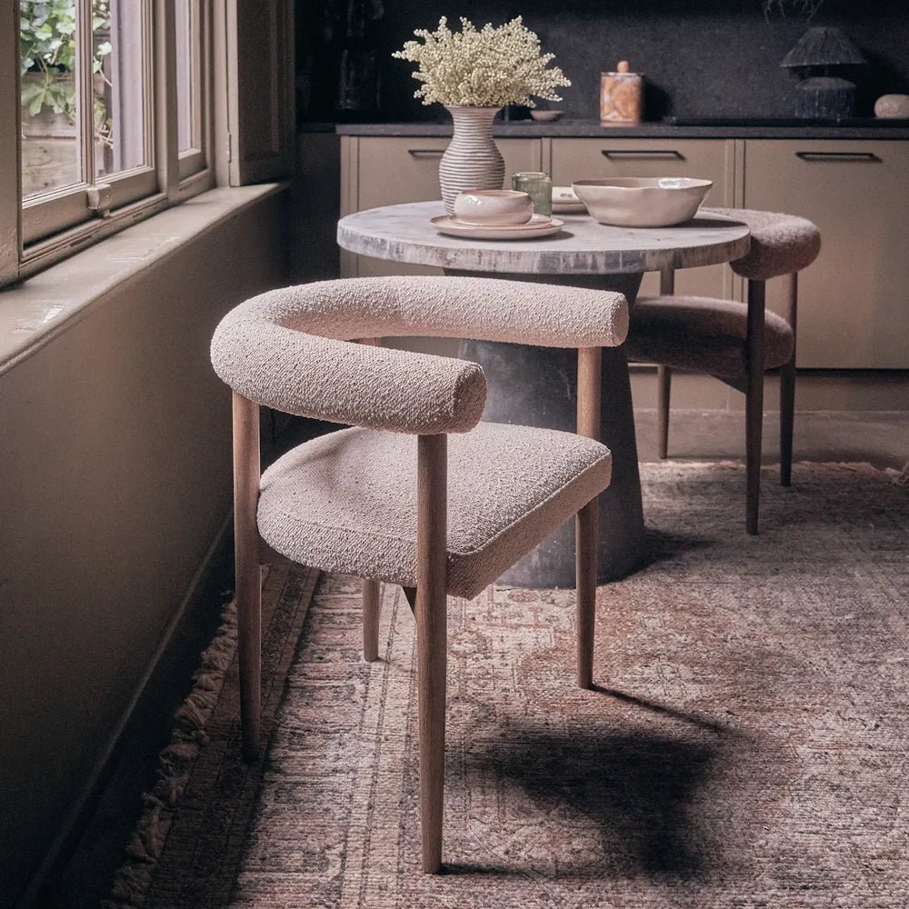 Harrington Boucle Dining Chair shows its curved cream backrest, open back and wooden frame, positioned beside a round dining table near a window.