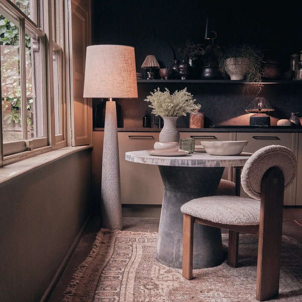 Haverford Polyresin Floor Lamp features a tall tapered textured base and pale fabric shade, standing beside a window in a softly styled dining area.