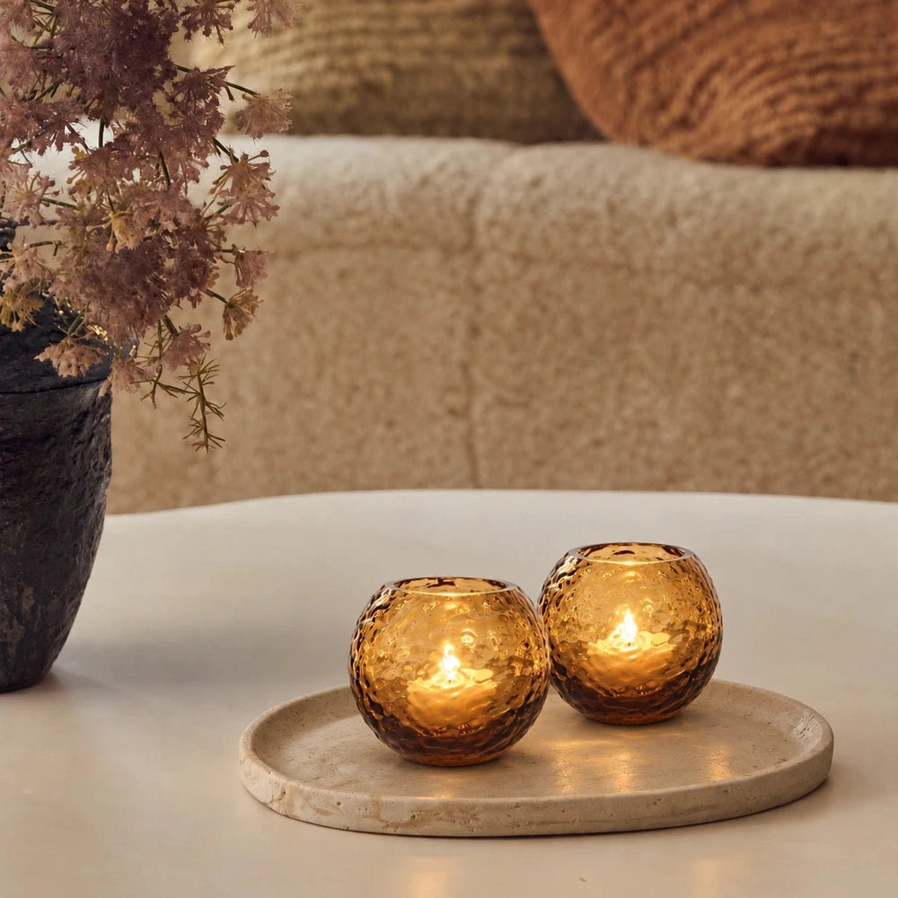Two rounded amber Hawick glass candle holders with dimpled surfaces glow on a pale oval tray, beside flowers and a cream sofa with rust cushions.