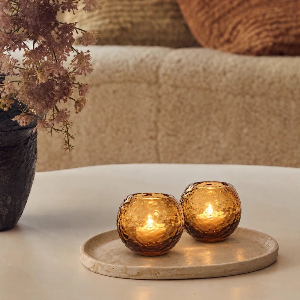 Two rounded amber Hawick glass candle holders with dimpled surfaces glow on a pale oval tray, beside flowers and a cream sofa with rust cushions.