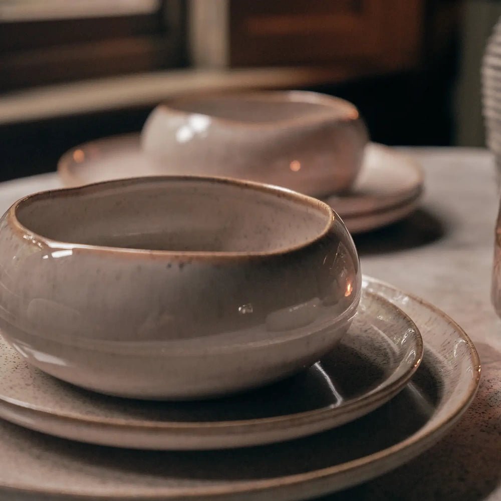 Hollister bowls sit on matching rounded plates, showing a glossy taupe glaze, softly irregular rims and speckled surfaces on a pale dining table.