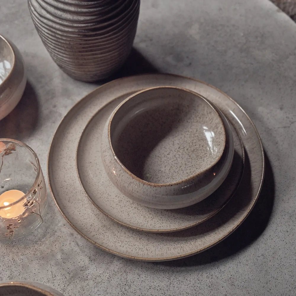 Hollister dinnerware features rounded taupe plates and a deep bowl with a softly speckled glaze, stacked on a stone tabletop beside a ribbed vase.