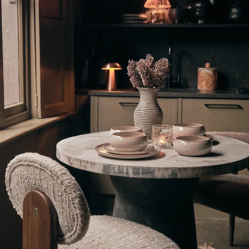 Hollister dinnerware creates a layered table setting on a round pale table, with textured chairs, a ribbed vase and candlelight in a dark dining room.