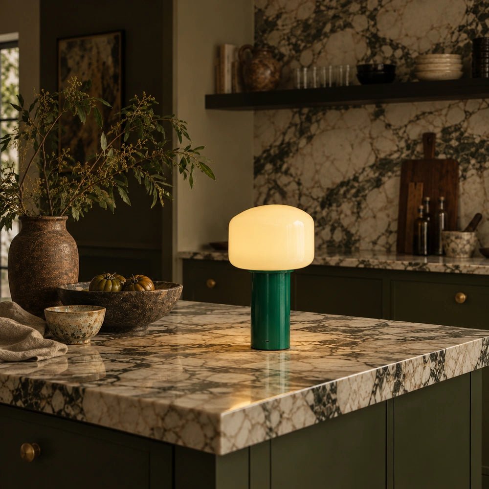 Green Hudson portable table lamp glowing on a veined stone kitchen island, surrounded by dark cabinetry, ceramics and leafy foliage in a moody kitchen.