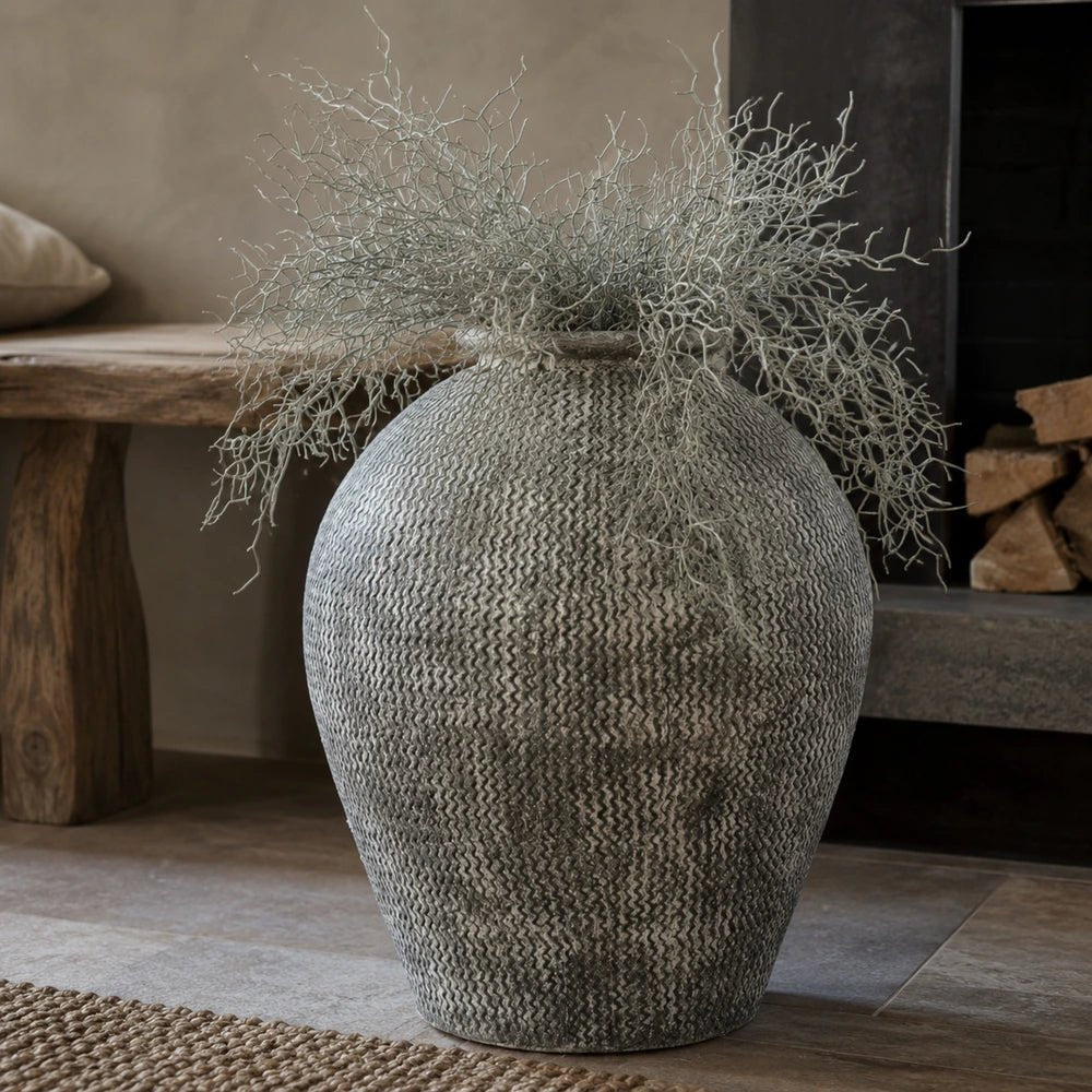 Immi Terracotta Vase with a rounded textured grey surface, styled with airy branching foliage on a tiled floor beside a dark fireplace and rustic wooden bench.