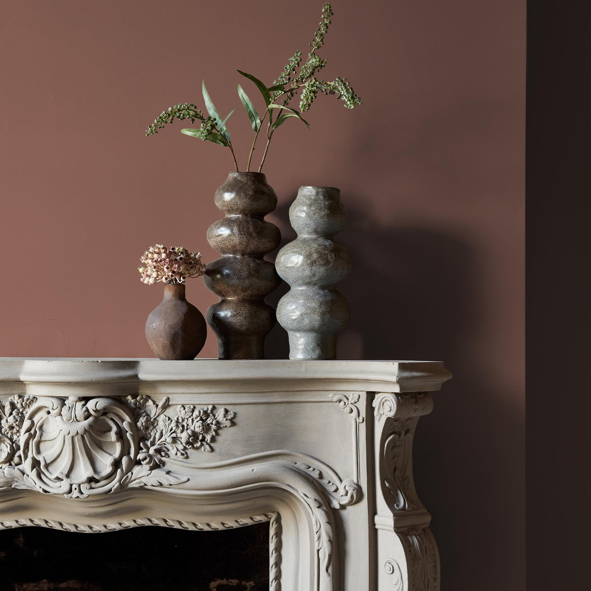Three ceramic vases sitting on an intricate fireplace mantle in front of a wall in a warm, terracotta paint colour.