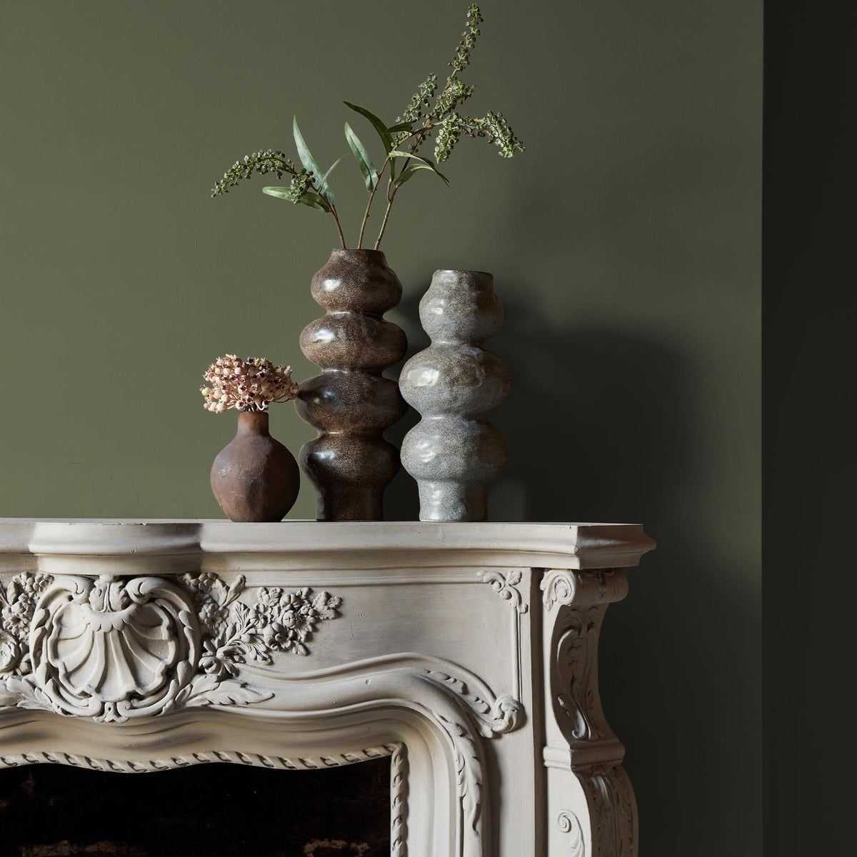 Three ceramic vases sitting on an intricate fireplace mantle in front of a wall in a warm, olive green paint.