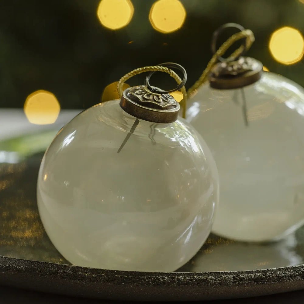 Kendal large opal bauble displays a rounded pearl white surface and dark metallic hanging cap in close view, resting beside another ornament on a decorative plate.