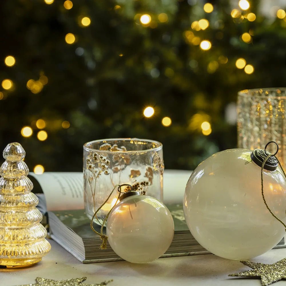 Kendal medium opal bauble with a round pearl white surface rests beside a larger ornament and patterned candleholder, with Christmas tree lights glowing behind.