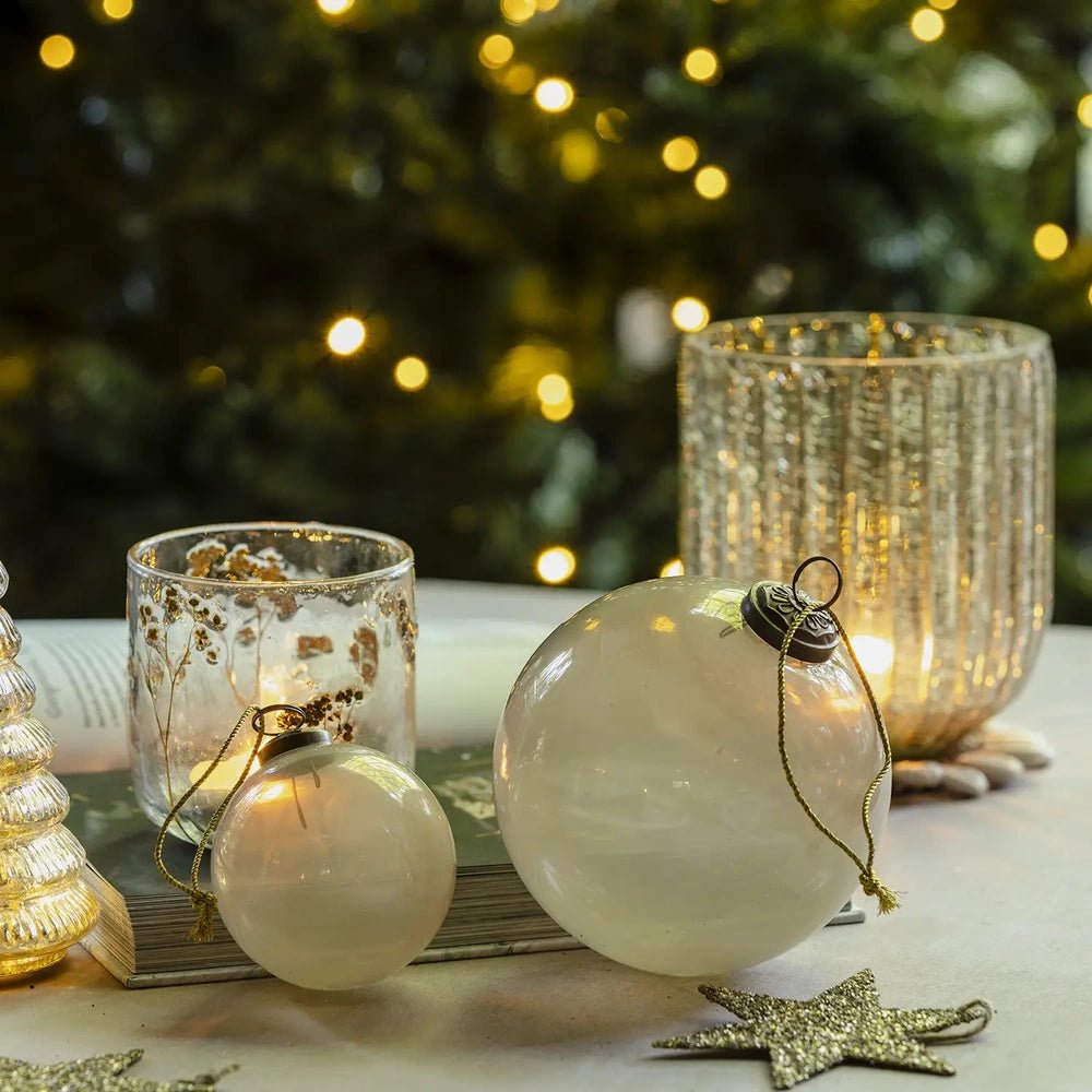 Kendal medium opal bauble displays a smooth creamy surface and dark hanging cap, arranged beside a larger ornament, glowing candleholder and ribbed glass lantern.