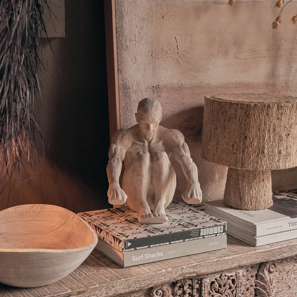 Lawrence Figurative Sculpture shows a pale seated figure on stacked books, arranged with sculptural vessels and a cylindrical lamp on a wooden console table.