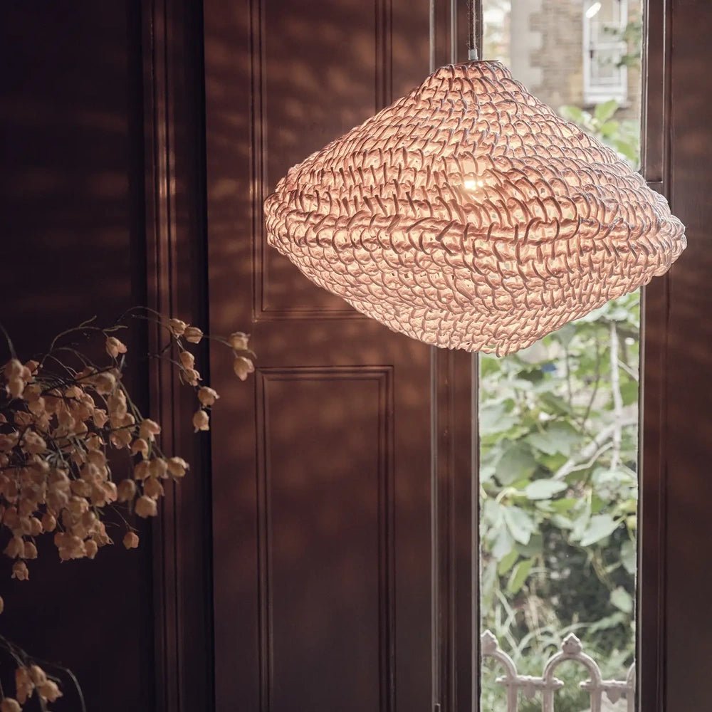Livington Paper Rope Pendant glows through an open woven shade, suspended beside a tall window above dried branches in a dark wood-panelled interior.