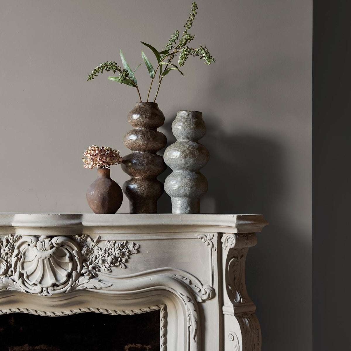 Three ceramic vases sitting on an intricate fireplace mantle in front of a wall in a soft greige paint colour.