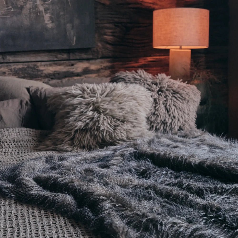 Malton Throw drapes across layered grey bedding with a long shaggy surface, accompanied by textured cushions and an orange-shaded lamp in a moody bedroom.