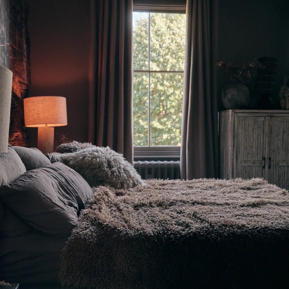 Mesa Faux Fur Throw brings a thick taupe shaggy texture to a bed, with soft cushions and an orange-shaded lamp beside a tall window.