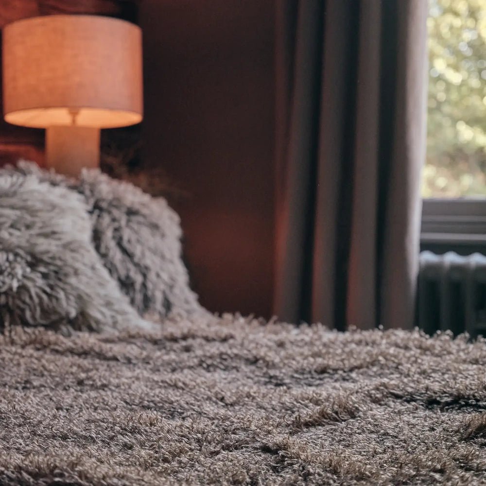 Mesa Faux Fur Throw displays dense taupe fibres across its shaggy surface, shown closely with textured cushions and a warm bedside lamp softly behind.