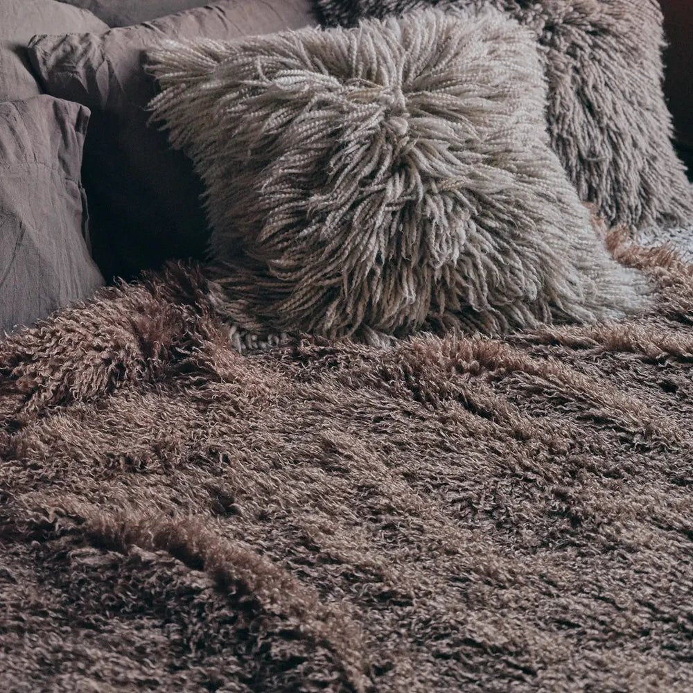 Mesa Faux Fur Throw covers a bed with a thick taupe shaggy texture, shown beside matching textured cushions and soft grey bedding in close-up.