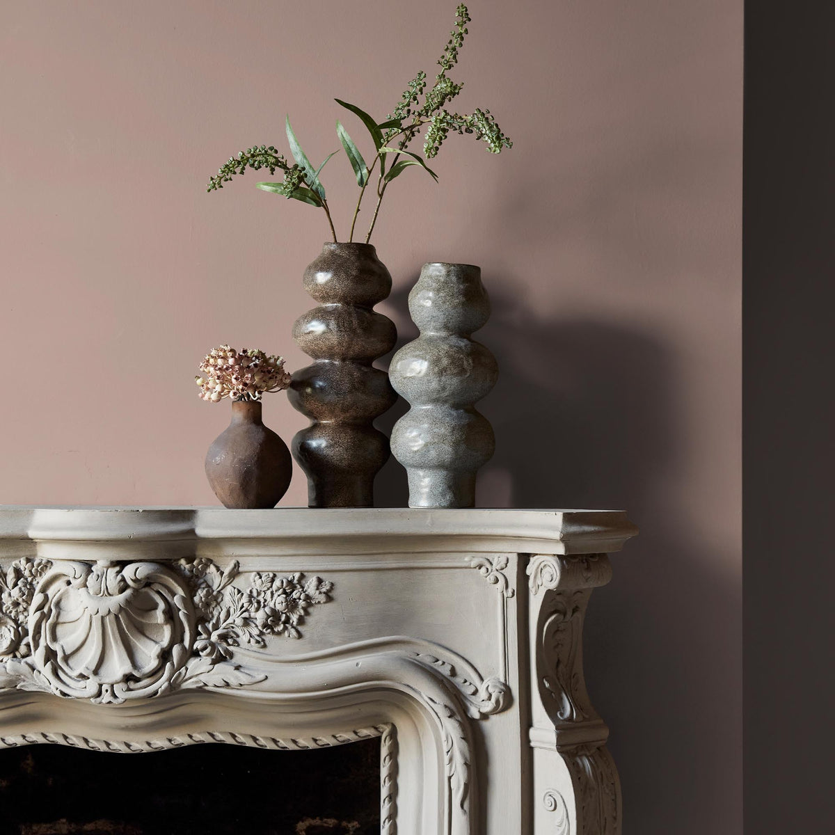 Three ceramic vases sitting on an intricate fireplace mantle in front of a wall in a warm, blush pink paint colour.