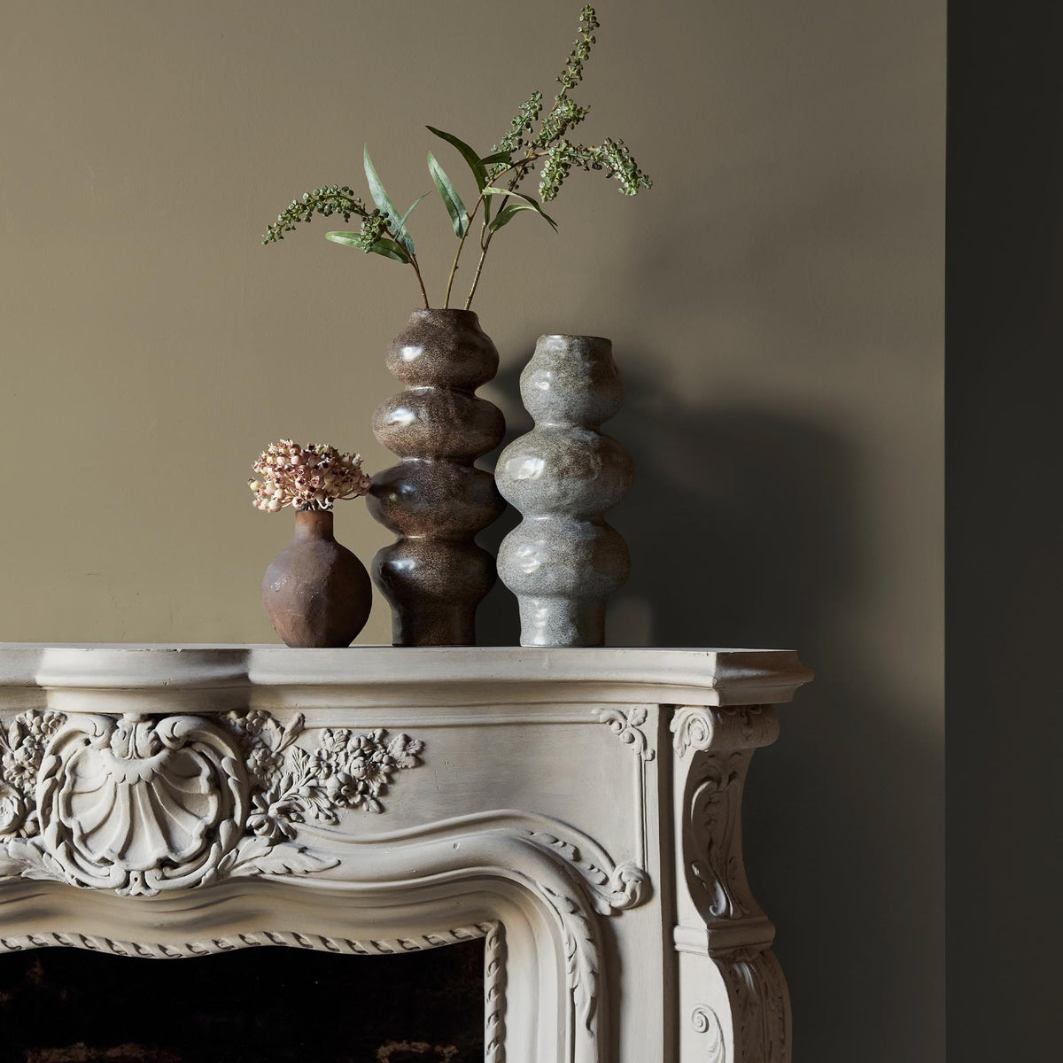 Three ceramic vases sitting on an intricate fireplace mantle in front of a wall in a neutral, sage green paint colour.