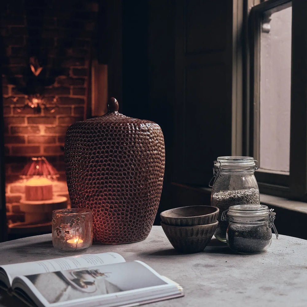 Nara Ceramic Jar in terracotta stands on a pale table beside glassware, bowls and candlelight, with its dimpled surface visible against a glowing fireplace.